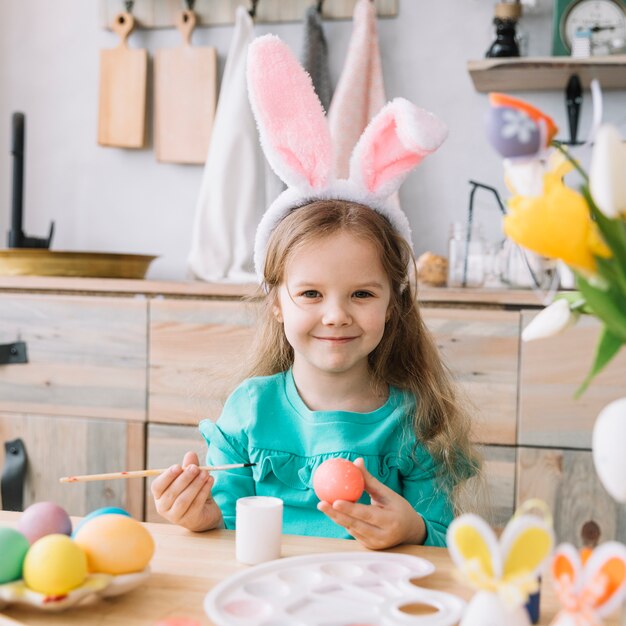 Linda chica en orejas de conejo pintando huevos para Pascua