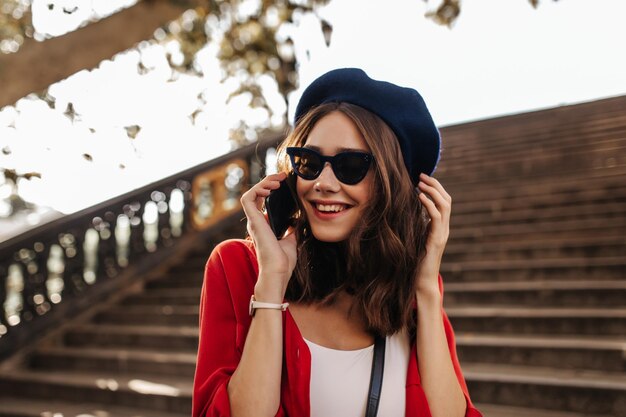 Linda chica pálida con cabello ondulado morena en boina gafas de sol de moda top blanco y camisa roja sonriendo y hablando por teléfono contra el fondo de las escaleras de la ciudad Otoño cálido y soleado