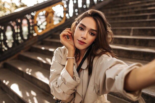 Linda chica pálida con maquillaje brillante y cabello oscuro haciendo selfie en gabardina beige y hermosa posando al aire libre