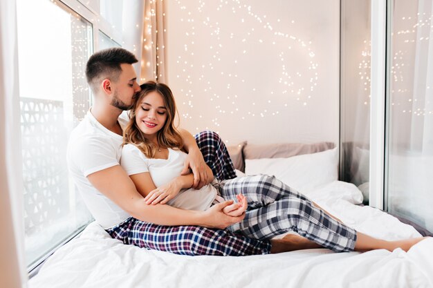 Linda chica en pantalones a cuadros acostada en la cama con su novio. Modelo masculino romántico besando el cabello de la esposa en la mañana.