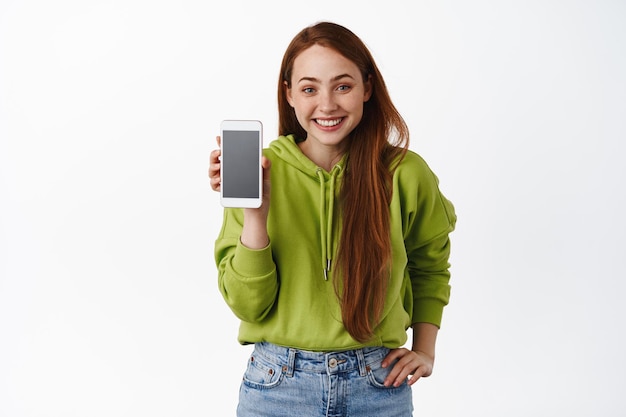 Linda chica pelirroja adolescente, muestra la aplicación del teléfono móvil y sonríe feliz, presenta la interfaz de la aplicación del teléfono inteligente, de pie contra el fondo blanco