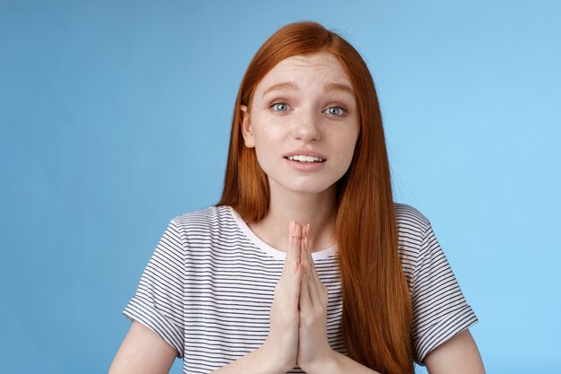 Linda chica pelirroja europea ojos azules peinado largo y recto de jengibre haciendo promesa rogándote que te ayudes a presionar las palmas juntas rezando di por favor diciendo necesito favor preocupado suplicante, fondo azul.