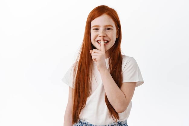 Linda chica pelirroja con pecas sonriendo con dientes y haciendo silencio, diciendo shhh contando un secreto, pidiendo estar en silencio, presionando el dedo en los labios y riendo, divirtiéndose, fondo blanco