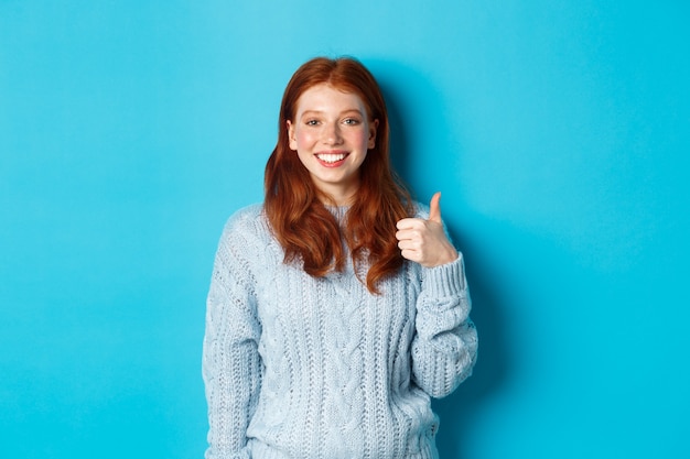 Linda chica pelirroja en suéter mostrando el pulgar hacia arriba, como y de acuerdo, sonriendo complacido, de pie contra el fondo azul