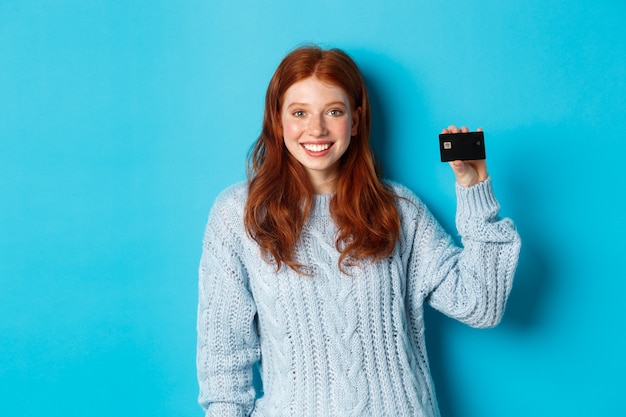 Linda chica pelirroja en suéter mostrando tarjeta de crédito, sonriendo a la cámara, de pie sobre fondo azul.