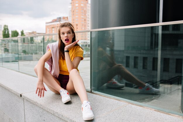 Linda chica de pelo castaño en zapatos blancos de moda sentado en el parapeto de piedra con expresión de cara de sorpresa