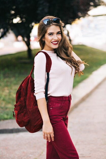 Linda chica con pelo largo está sonriendo en el parque de la ciudad. Ella tiene color marsala en sus cosas. Ella parece disfrutar.