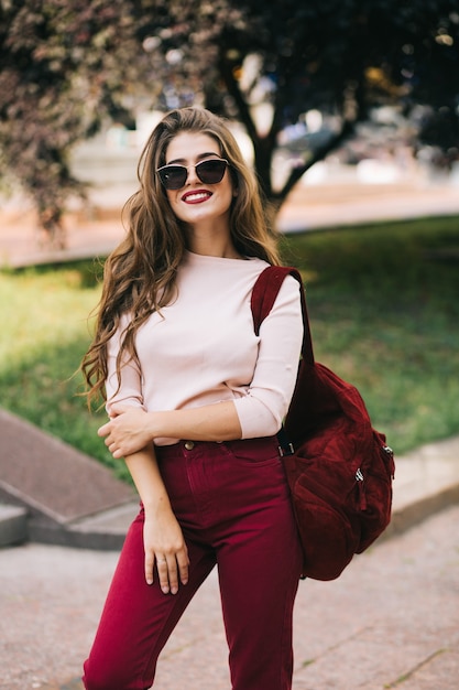 Linda chica con pelo largo en gafas de sol con bolso y pantalones vinoso está sonriendo en el parque de la ciudad.