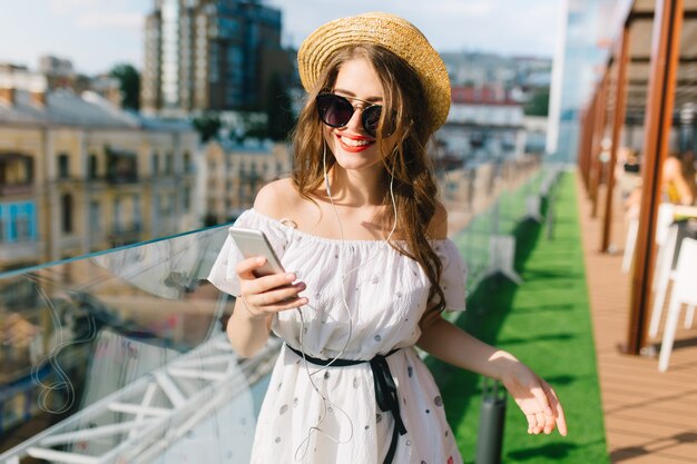 Linda chica con el pelo largo con gafas de sol está de pie en la terraza. Lleva un vestido blanco con hombros descubiertos, lápiz labial rojo y sombrero. Ella escucha música a través de auriculares.