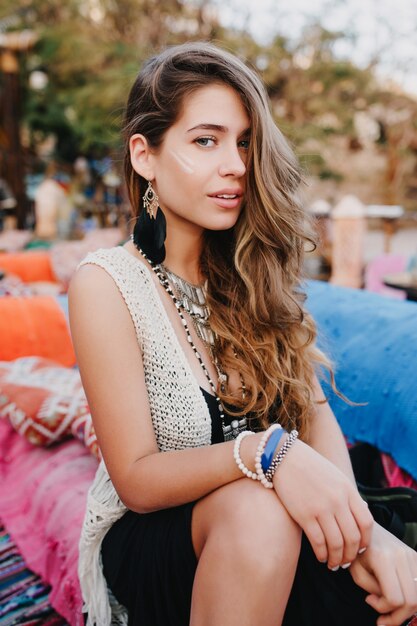 Linda chica de pelo largo en pulseras y aretes de moda posando afuera mientras descansa en la fiesta del parque