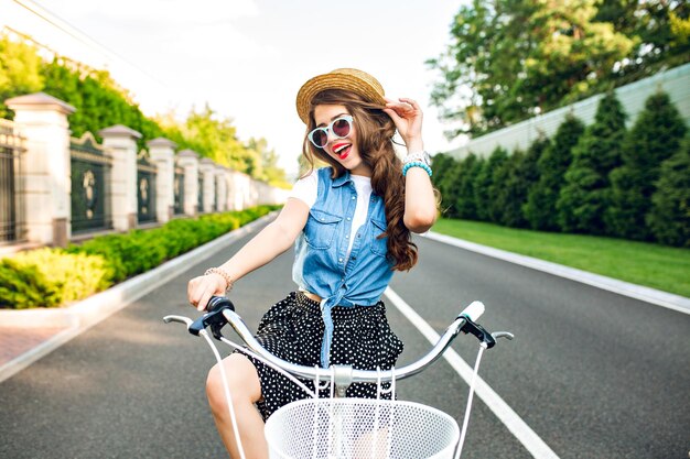 Linda chica con pelo largo y rizado en gafas de sol va en bicicleta a la cámara en la carretera. Lleva falda larga, jubón, sombrero. Ella luce feliz.