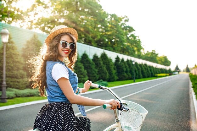 Linda chica con el pelo largo y rizado en gafas de sol va en bicicleta por la carretera. Lleva falda larga, jubón, sombrero. Ella se ve feliz bajo el sol.