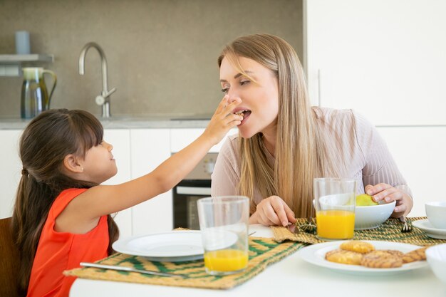 Linda chica de pelo negro dando galleta a su mamá para probar y morder, desayunando con su familia