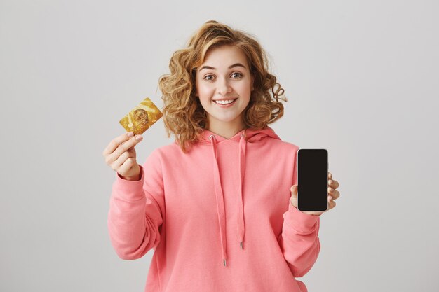 Linda chica de pelo rizado mostrando tarjeta de crédito dorada y pantalla de teléfono móvil