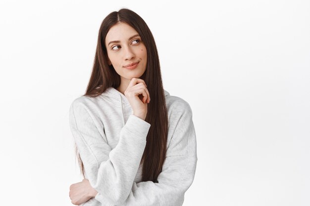 Linda chica pensativa con cabello largo mirando a un lado y sonriendo mientras piensa, de pie pensativo, tomando decisiones, imagina algo interesante, pared blanca