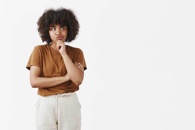 Linda chica pensativa concentrada con peinado afro en camiseta marrón y pantalones sonriendo mirando a la derecha mientras piensa con dudas