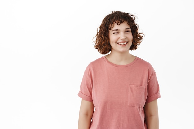 Linda chica positiva, guiña un ojo y te sonríe, muestra una sonrisa de dientes blancos perfectos, insinuando algo interesante, de pie feliz en blanco.