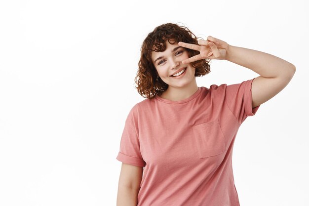 Linda chica positiva sonriendo feliz, mostrando paz kawaii v-sign y mirando a la cámara, parada encantadora contra fondo blanco en camiseta.