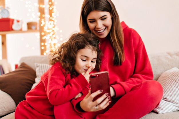 Linda chica rizada con smartphone con madre. Mamá joven sonriente escalofriante con hija preadolescente en el sofá.