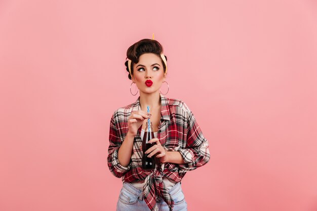 Linda chica rosa pensando en algo. Mujer elegante en camisa a cuadros sosteniendo una botella de bebida.