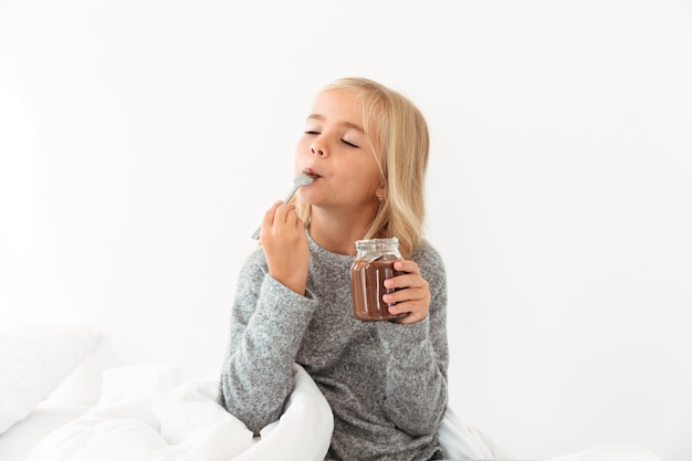 Linda chica rubia disfrutando chocolate avellana con los ojos cerrados mientras está sentado en la cama