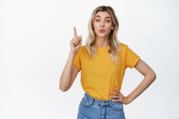 Linda chica rubia frunciendo los labios mostrando algo apuntando con el dedo a un anuncio o banner de venta de pie en camiseta amarilla y jeans fondo blanco.