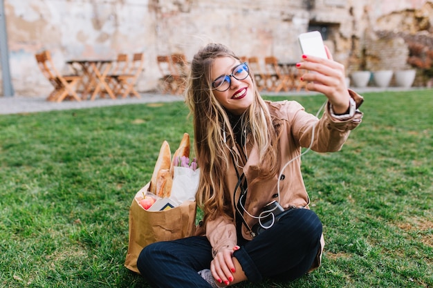 Linda chica rubia con gafas haciendo selfie con la mano sentada en la hierba verde en el parque. encantadora mujer joven descansando después de comprar y hacer fotos para el perfil de instagram con las piernas cruzadas