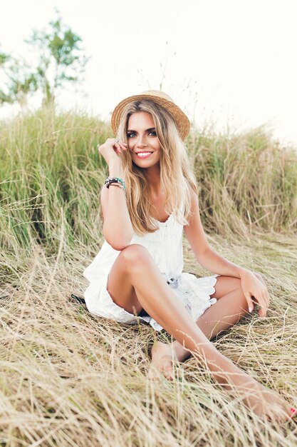 Linda chica rubia de longitud completa con cabello largo está posando para la cámara sobre pasto seco. Lleva sombrero, vestido blanco y sonríe a la cámara.