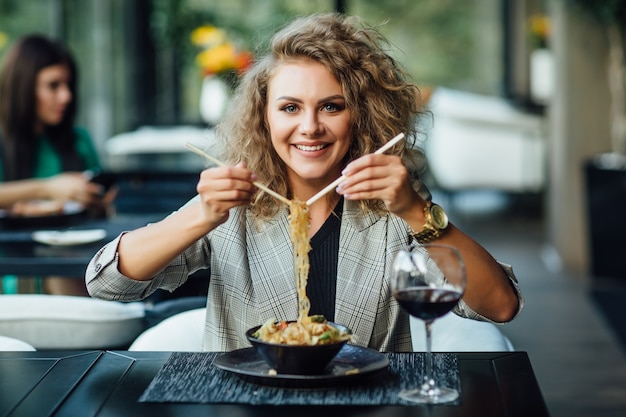 Linda chica rubia con palillos en un restaurante en barco. Chica intenta salmón con arroz. Chica come electrodomésticos. La chica de la chaqueta después del trabajo come.