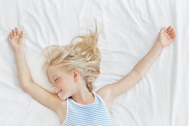 Linda chica rubia pequeña en camiseta de marinero durmiendo en una cama cómoda en ropa de cama blanca, sonriendo mientras tiene sueños agradables. Niña siente relajación en la cama estar cansado después de largos juegos