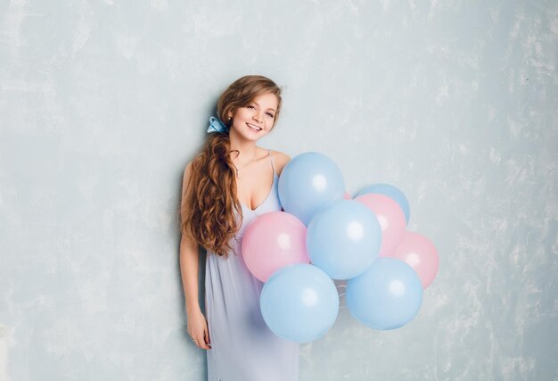 Linda chica rubia de pie en un estudio, sonriendo y sosteniendo globos azules y rosados. Viste de color azul claro y tiene el pelo largo trenzado y ondulado con una cinta azul.