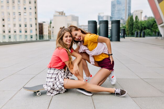 Linda chica rubia riendo con piernas largas sentada en patineta cerca de su hermana morena en camisa amarilla