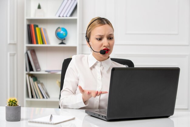 Linda chica rubia de servicio al cliente con camisa blanca con computadora portátil y auriculares molesta en una videollamada