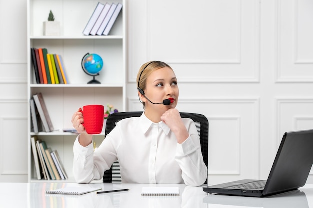 Linda chica rubia de servicio al cliente con camisa blanca con computadora portátil y auriculares sosteniendo una taza roja