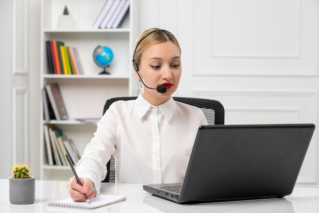 Linda chica rubia de servicio al cliente con camisa blanca con computadora portátil y auriculares tomando notas