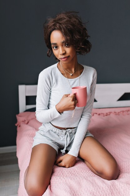 Linda chica sentada en la cama, tomando una taza de café, bebiendo té en la habitación con pared gris. Tiene el pelo corto y rizado. El uso de pantalones cortos de color gris claro, camiseta de manga larga.