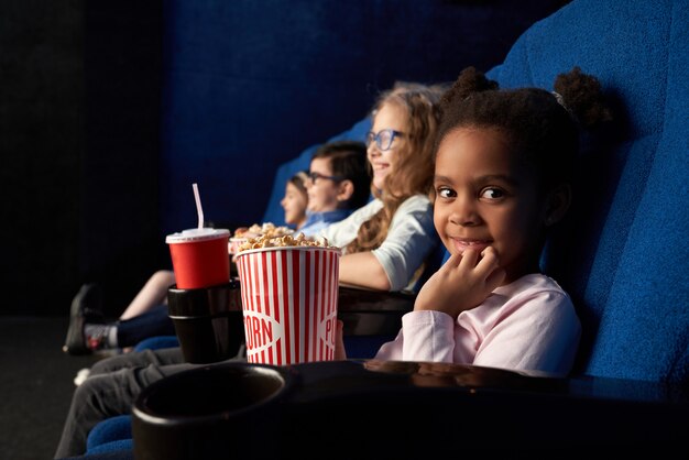 Linda chica sentada en el cine con amigos, mirando a cámara.