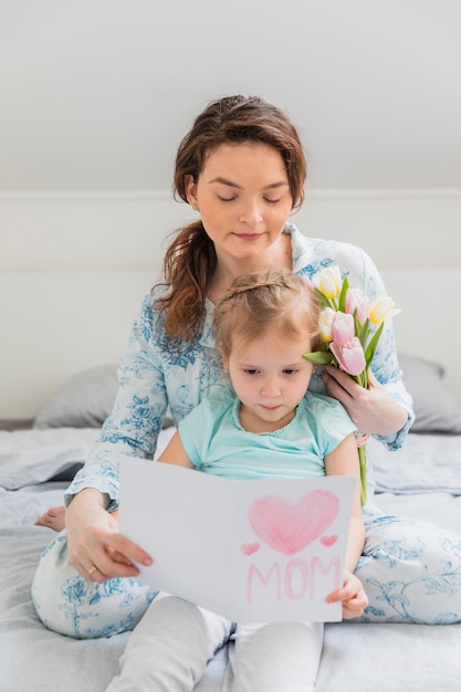 Linda chica sentada con su madre leyendo la tarjeta de felicitación en la cama