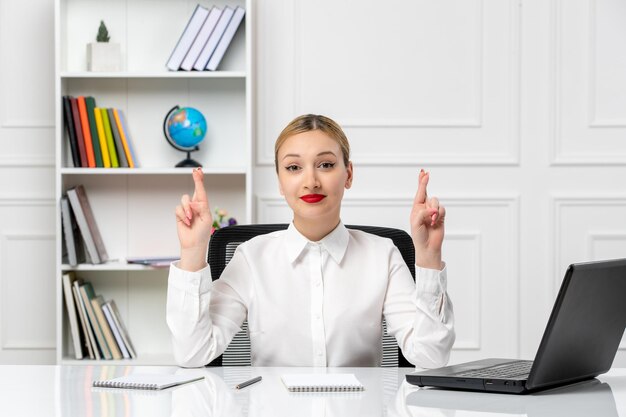 Linda chica de servicio al cliente en camisa blanca con lápiz labial rojo y computadora portátil cruzando los dedos