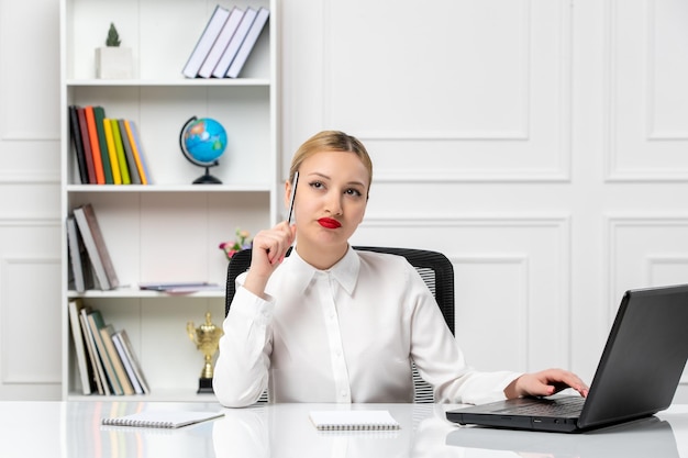 Linda chica de servicio al cliente con camisa blanca con lápiz labial rojo y computadora portátil pensando con bolígrafo en la sien