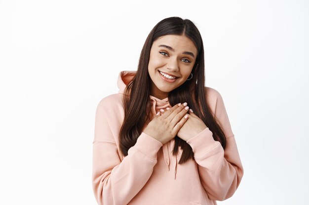 Linda chica sincera recibe un regalo, cogidos de la mano en el corazón y sonriendo complacida, da las gracias, expresa gratitud, de pie en una sudadera con capucha contra la pared blanca