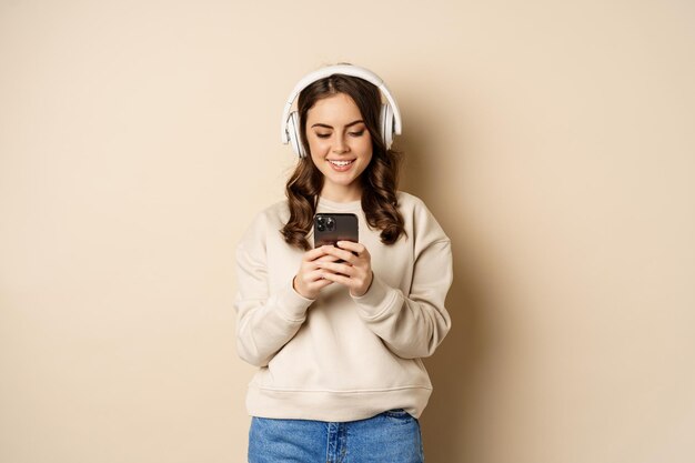 Linda chica sonriente con auriculares, mirando el teléfono móvil, escuchando música o podcast, de pie sobre un fondo beige