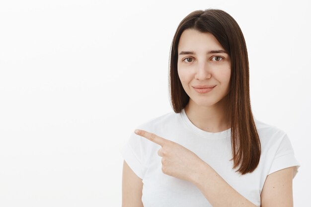 Linda chica sonriente con cabello castaño y ojos en camiseta blanca sonriendo tiernamente mientras apunta a la izquierda con el dedo índice