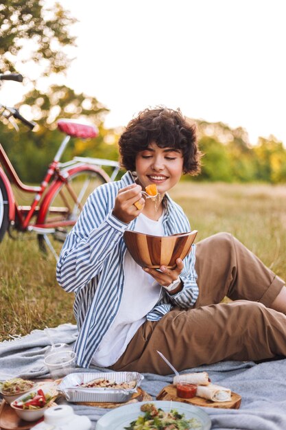 Linda chica sonriente con camisa a rayas sentada en una manta comiendo ensalada en un picnic con bicicleta en el fondo del parque