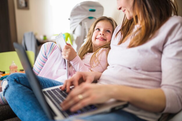 Linda chica sonriente mirando a su madre usando la computadora portátil