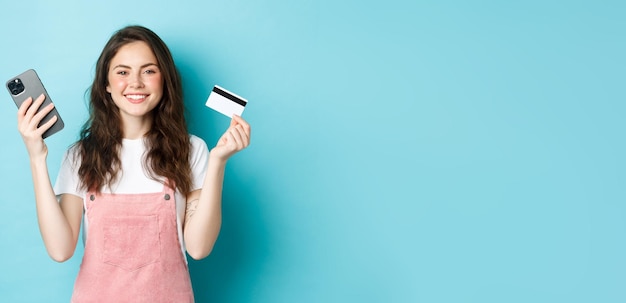 Foto gratuita linda chica sonriente que muestra una tarjeta de crédito de plástico y un teléfono inteligente pide algo en la tienda a través de la tienda de aplicaciones móviles en línea o paga un pedido de fondo azul