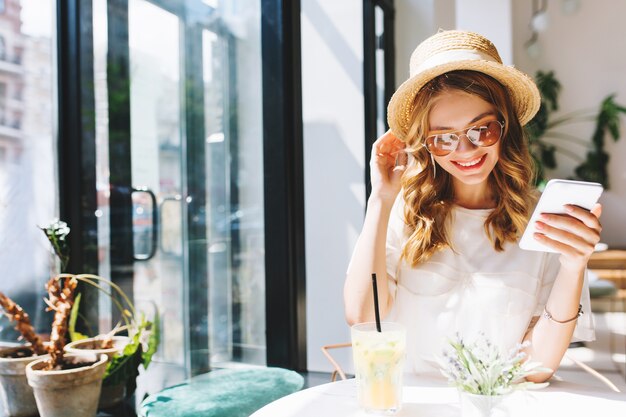 Linda chica sonriente con sombrero de paja relajándose en la cafetería al lado de la puerta de cristal sosteniendo el smartphone en la mano