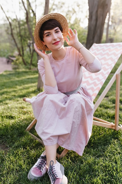 Linda chica sonriente en traje de moda disfrutando de fin de semana y hermosas vistas de la naturaleza, sentada en una silla de jardín