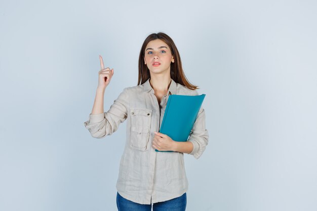 Linda chica sosteniendo la carpeta, apuntando hacia arriba en la camisa y mirando inteligente