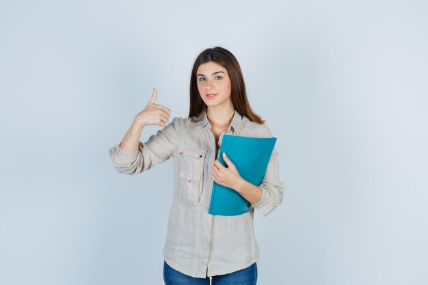 Linda chica sosteniendo la carpeta, mostrando el pulgar hacia arriba en la camisa y mirando complacido